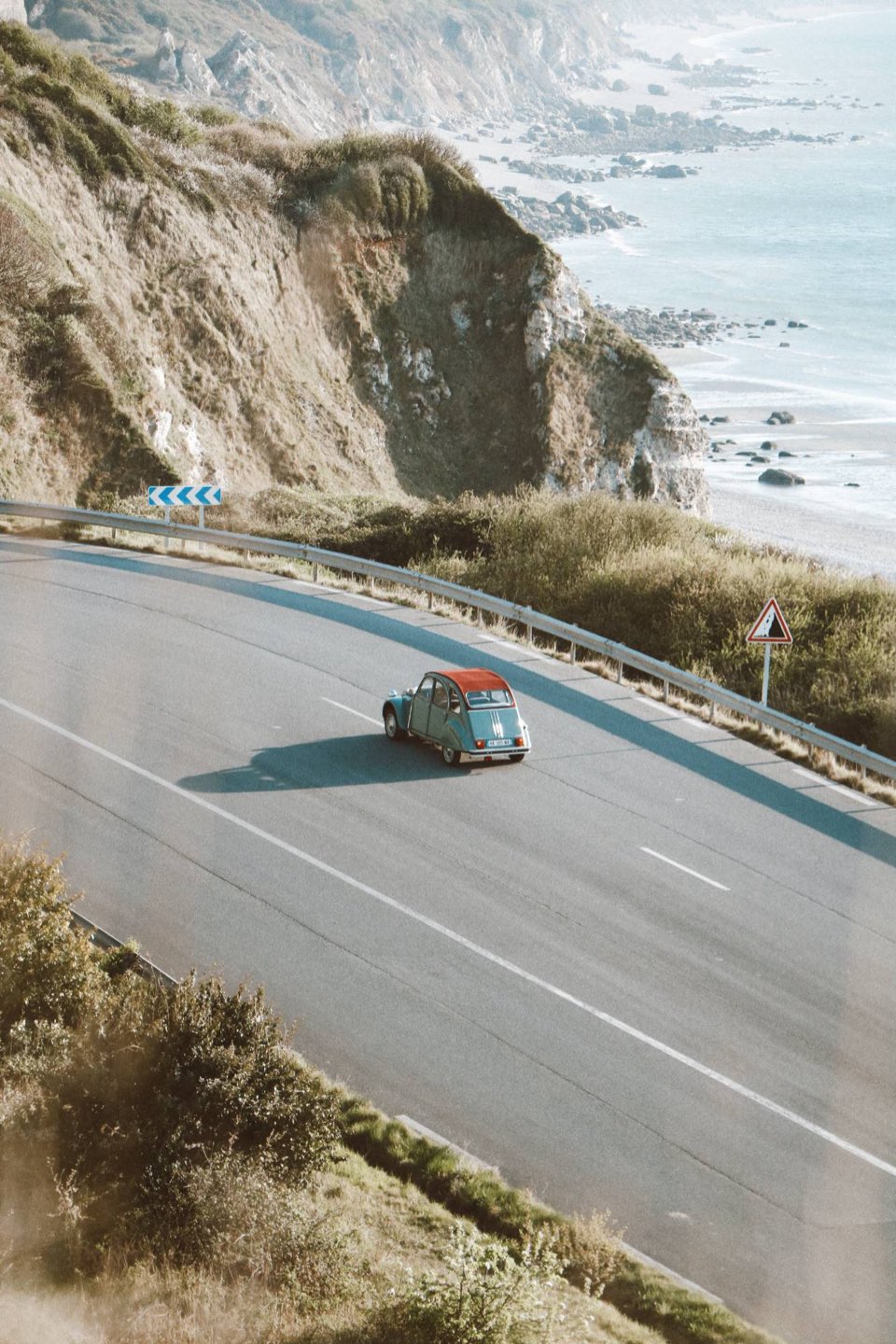 2CV turquoise sur une route en lacets surplombant la mer.