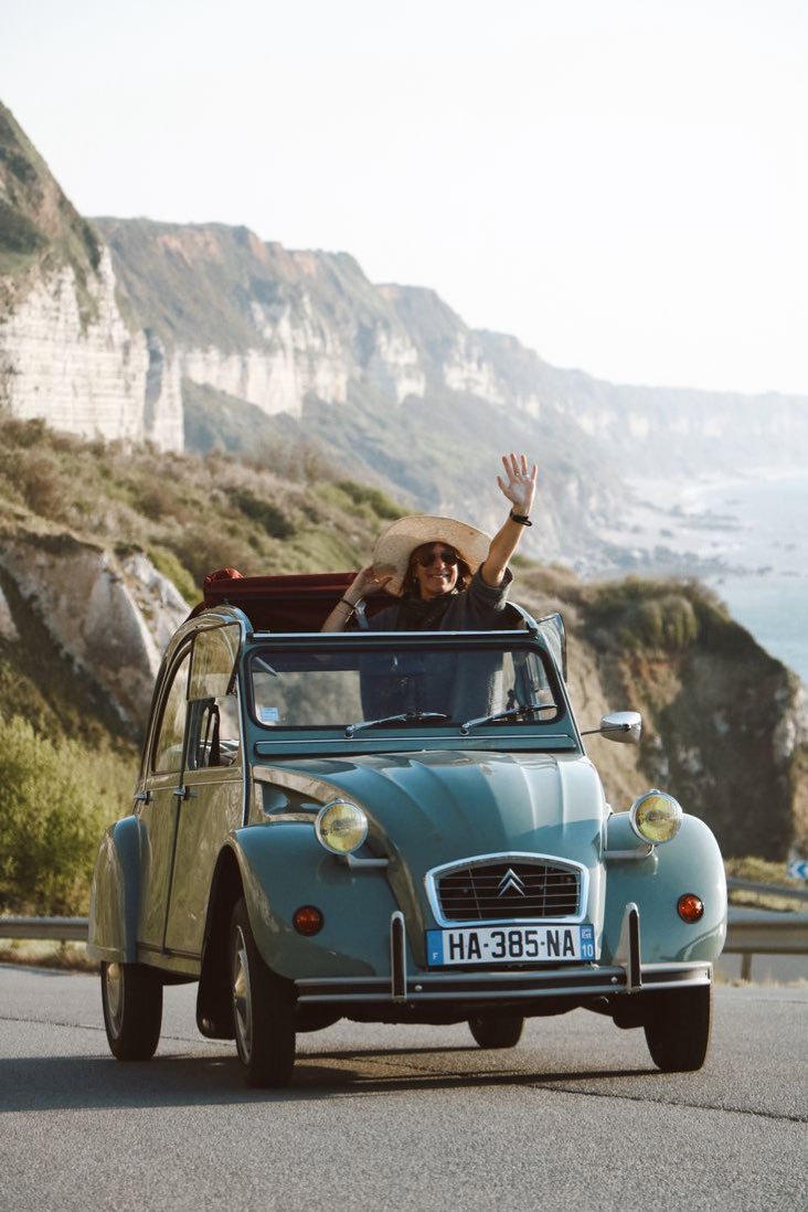 2CV bleue sur une route côtière, une passagère salue depuis le toit ouvrant, falaises en arrière-plan.